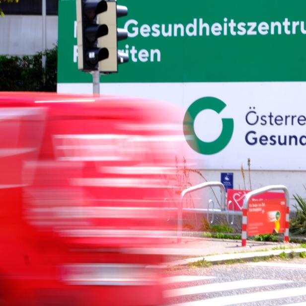 Ein Schild der Österreichischen Gesundheitskasse (ÖGK) mit einem vorbeifahrenden roten Lieferwagen.