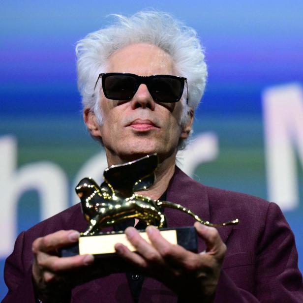 Mann mit weißem Haar und dunkler Sonnenbrille hält die goldene Statue eines geflügelten Löwen in seinen Händen