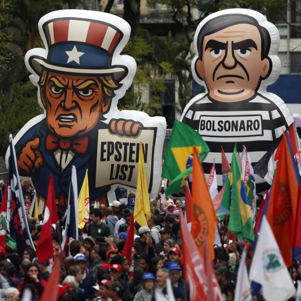 Man sieht einen Demozug mit Balloonfiguren von Bolsonaro in einem Gefängnisoutfit und Trump, der ein Stück Papier mit der Aufschrift "Epstein List" in der Hand hält.