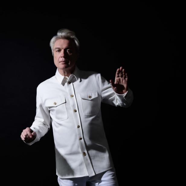 Ex-Talking-Heads-Chef David Byrne, 73 Mann in Halbschatten und langem weißen Hemd hebt eine Hand