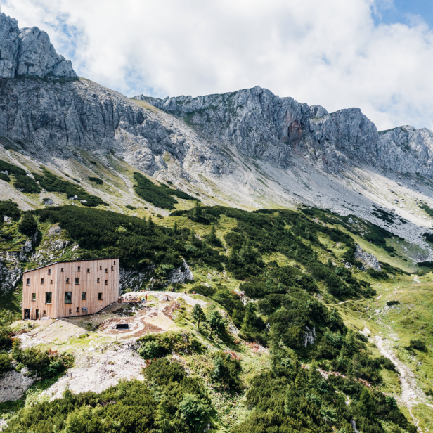 Ein modernes Holzgebäude steht inmitten einer alpinen Landschaft mit grünen Hängen und steilen Felswänden im Hintergrund.