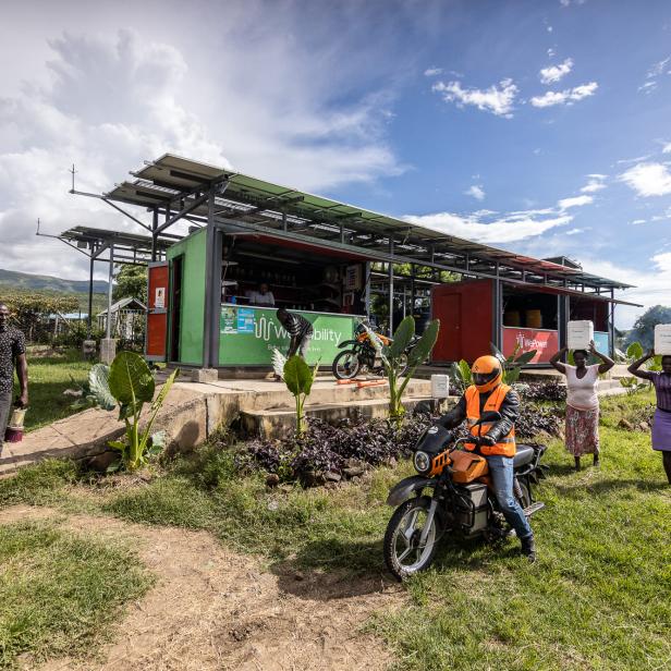 Grünes Containergebäude mit Solarpanelen auf dem Dach, davor eine Person auf einem Motorrad mit orangefarbenem Helm und Weste, zwei Frauen tragen Behälter auf dem Kopf, ländliche Umgebung mit Gras und Bäumen.