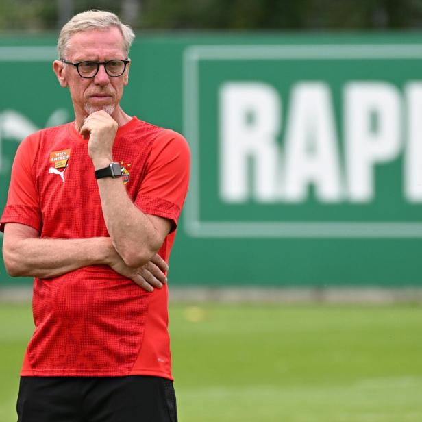 Rapid-Trainer Peter Stöger steht auf einem Fußballplatz, trägt rotes T-Shirt, im Hintergrund ein Rapid-Schriftzug