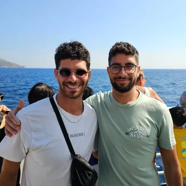 Die Brüder Ilay und Evyatar stehen, Arm in Arm, lachend am Meer. 
