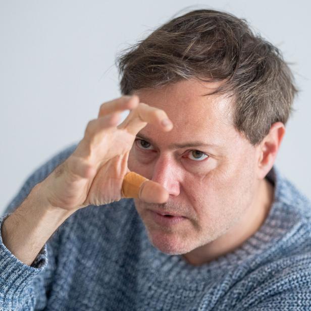 Festwochen-Intendant Milo Rau Mann in blauem Pullover hält seine rechte Hand mit ausgestreckten Fingern, als wollte er etwas packen, vors Gesicht