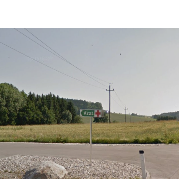 Ein Wegweiser mit der Aufschrift „Arzt“ steht an einer Landstraße vor Feldern und Wiesen am Rand von St. Marienkirchen am Hausruck.