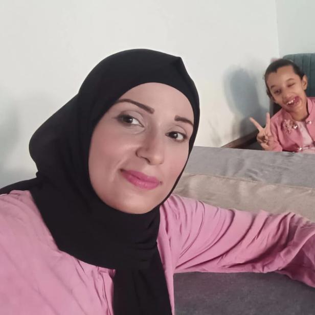 Eine Frau in Kopftuch posiert vor einem Esstisch für ein Selfie, am anderen Ende des Tische sitzen zwei Mädchen.