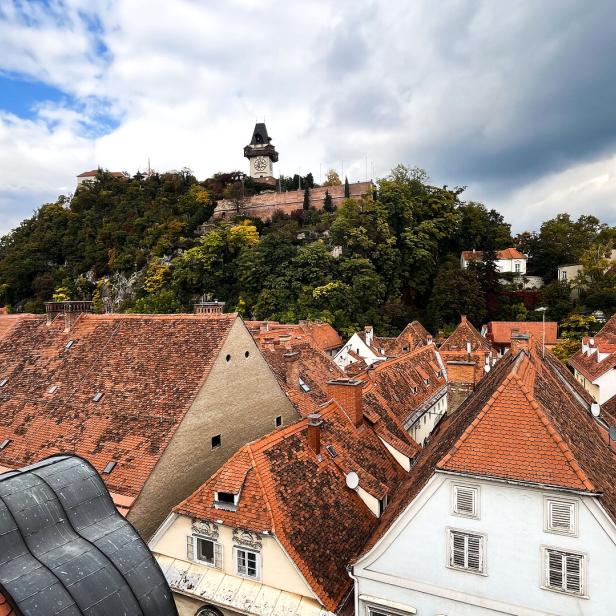 Die Dächer von Graz und der Uhrturm am Schlossberg. Der Himmel ist bewölkt.