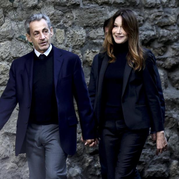 Nicolas Sarkozy und Carla Bruni gehen händchenhaltend vor einer Steinmauer entlang.