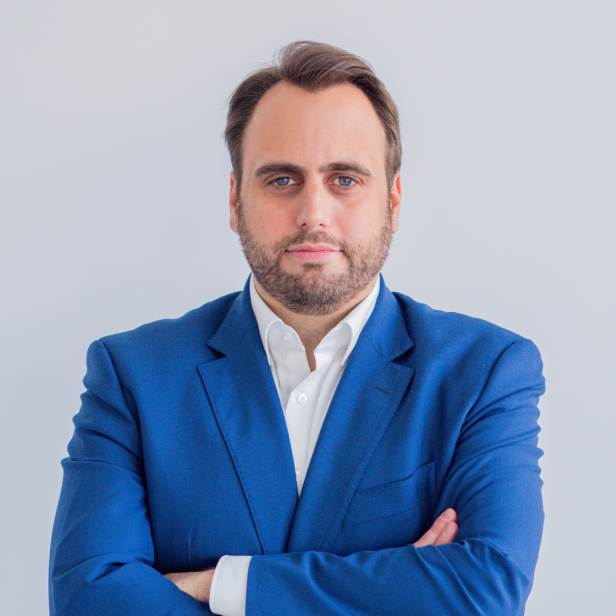 Mann mit blauem Sakko und weißem Hemd steht mit verschränkten Armen vor hellem Hintergrund.