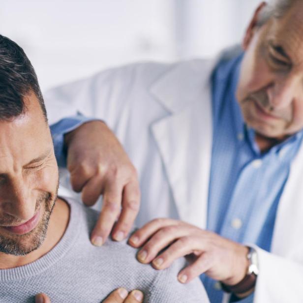 Das Bild zeigt eine Behandlungsszene, bei der ein Arzt einen Patienten untersucht, dessen Schulter schmerzt.