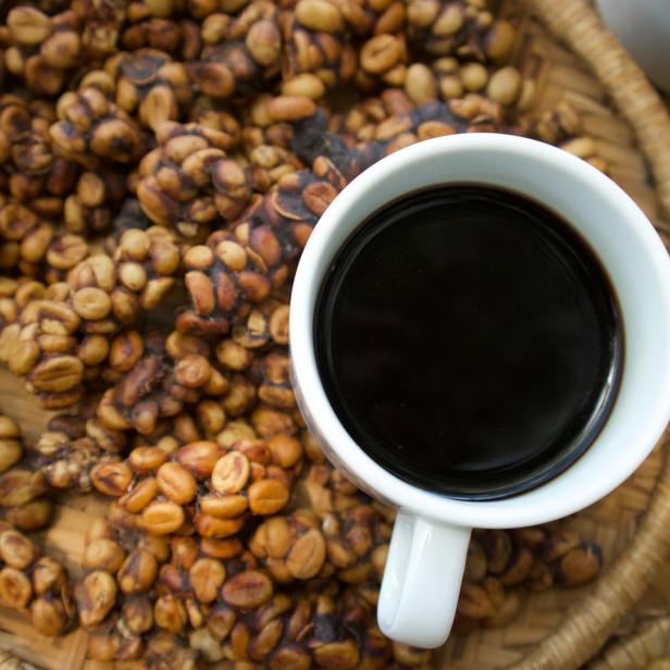 Das Bild zeigt eine Tasse mit schwarzem Kaffe auf einem Teller voller Kaffeebohnen. Es sind jene Bohnen, die von einer speziellen Schleichkatzenart gefressen und verdaut werden.