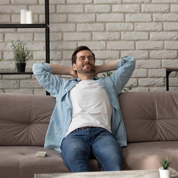 Person lehnt entspannt auf einem grauen Sofa, trägt blaue Jeans und hellblaue Hemdjacke, Hände hinter dem Kopf verschränkt./ Durch kontrollierte Wohnraumlüftung Schimmel vermeiden: air-plus Lüftungstechnik GmbH sorgt für saubere Luft, Energieeffizienz und Werterhalt von Immobilien.