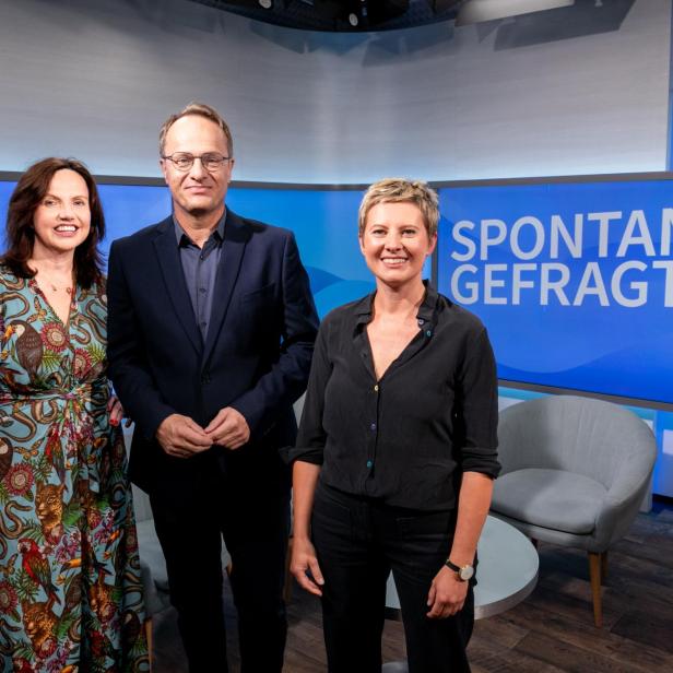 Drei Personen stehen vor einem blauen Bildschirm mit der Aufschrift 'SPONTAN GEFRAGT' in einem Fernsehstudio.