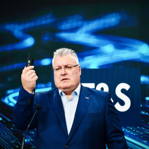 Michael Mertin hält eine Fernbedienung mit seiner rechten Hand hoch. Hinter ihm ist eine Vidi-Wall mit dunkelblauen und hellblauen Kabeln und Elektronik darauf.