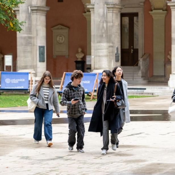 Gruppe von fünf Personen, die auf einem gepflasterten Platz vor einem historischen Gebäude mit Arkaden und Statuen (Arkadenhof der Uni Wien) gehen.