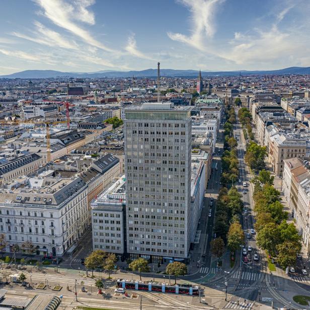 Luftaufnahme des Ringturms, eines hohen, rechteckigen Bürogebäudes, umgeben von Straßen, historischen Gebäuden und mehreren Straßenbahnen in Wien.
