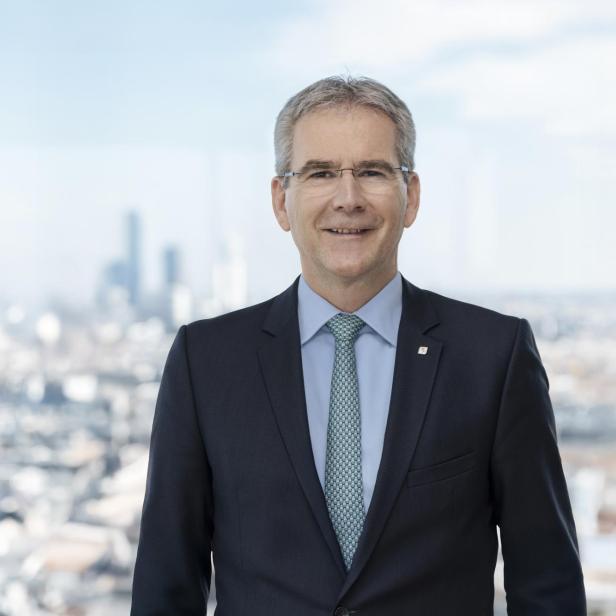 Mann in dunklem Anzug mit hellblauem Hemd und grün-blau gemusterter Krawatte vor unscharfer Stadtlandschaft.