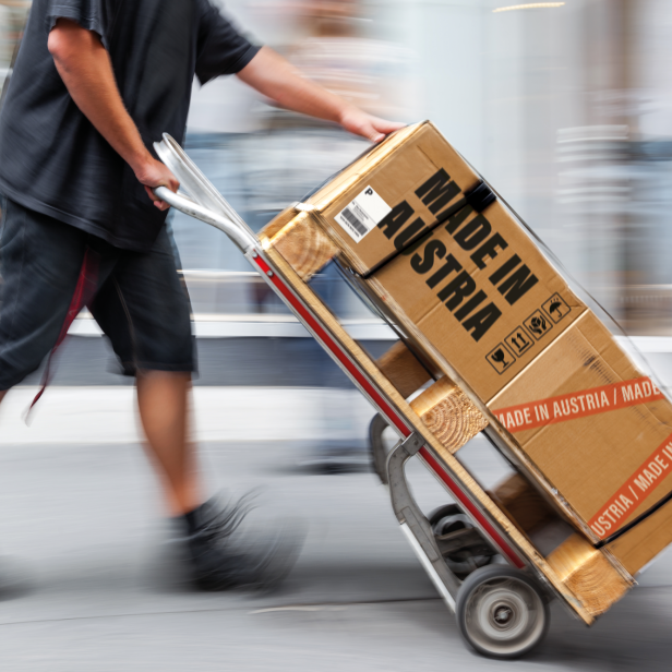 Person in schwarzer Kleidung schiebt Sackkarre mit zwei Kartons, auf denen 'MADE IN AUSTRIA' steht.