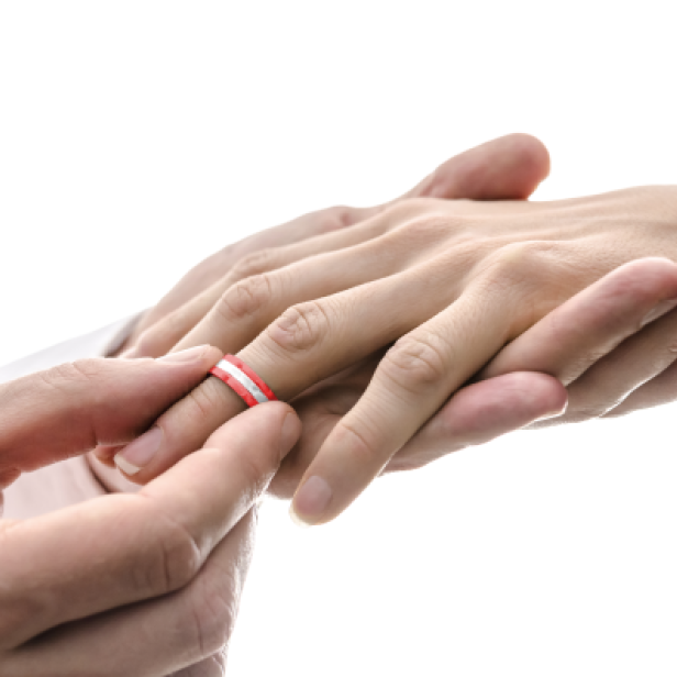 Zwei Hände, eine legt einen Ring mit rotem und weißem Muster auf den Ringfinger der anderen Hand.