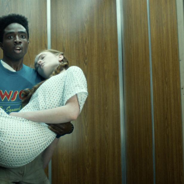 Junger Mann mit erschrockenem Gesicht trägt ein bewusstloses Mädchen und einen Kassettenrecorder vor holzvertäfeltem Hintergrund.