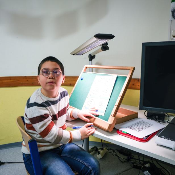 Ein Kind sitzt an einem Schreibtisch in einem Klassenraum und arbeitet an einer geneigten Lern- oder Lesetafel. Auf dem Tisch stehen weitere Lernmaterialien und ein großer Monitor.