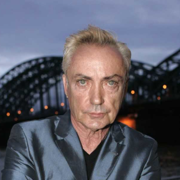 Blondierter Mann in silbergrauem Anzug blickt in die Kamera, im Hintergrund eine beleuchtete Brücke in der Abenddämmerung