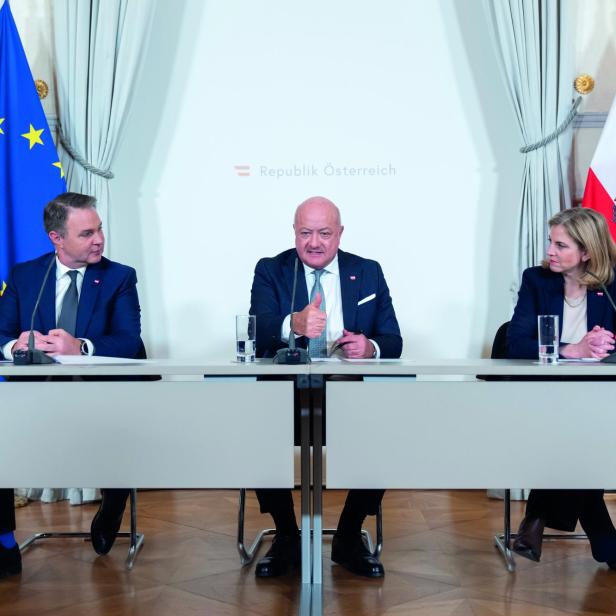 Andreas Babler, Christian Stocker und Beate Meinl Reisinger sitzen an zwei nebeneinanderstehenden Tischen, im Hintergrund je zwei österreichische und europäische Flaggen.