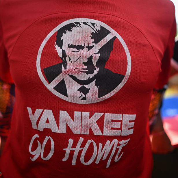Eine Person trägt ein rotes Shirt mit der Aufschrift "Yankee Go Home" und dem durchgestrichenen Konterfei von Donald Trump, dahinter eine Venezuela-Flagge