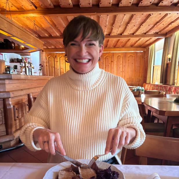 Astrid Steharnig-Staudinger sitzt in einem leeren Wirtshaus mit Holzvertäfelung und isst einen Schweinsbraten