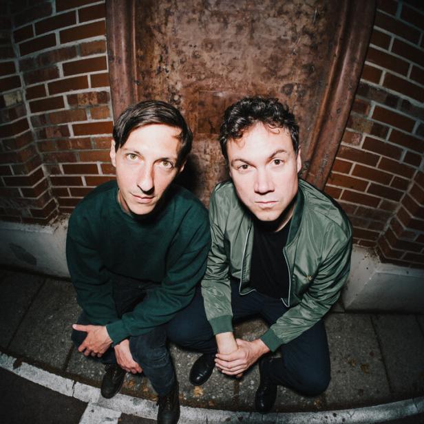 Zwei junge Männer in einer Art Keller blicken mit ernsten Gesichtern in die Fischaugen-Kamera 