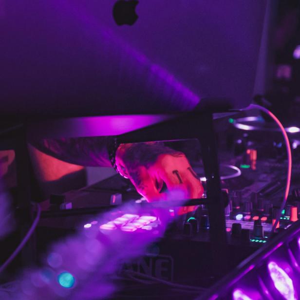 Ein DJ mit Tätowierung bedient ein Pioneer-Mischpult unter violettem Licht, ein Laptop steht daneben.
