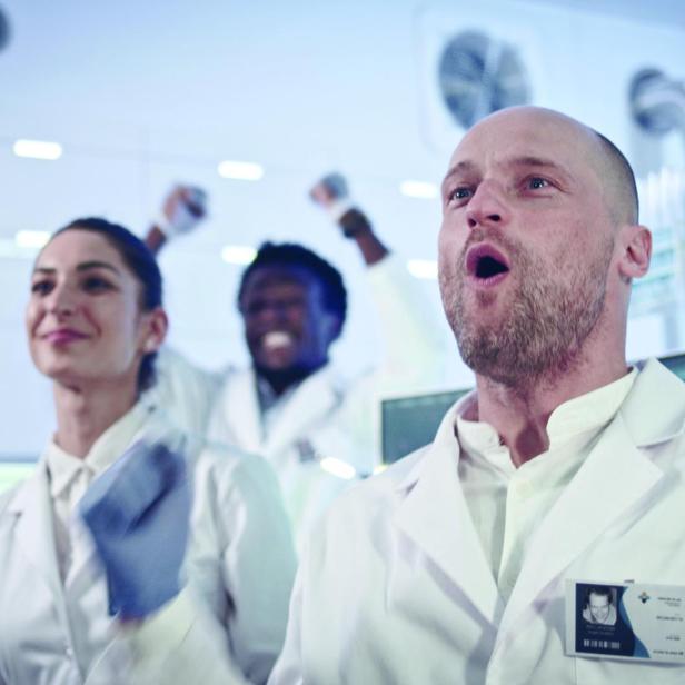 Gruppe von Wissenschaftlern in weißen Laborkitteln jubelt in einem Labor mit technischen Geräten im Hintergrund.