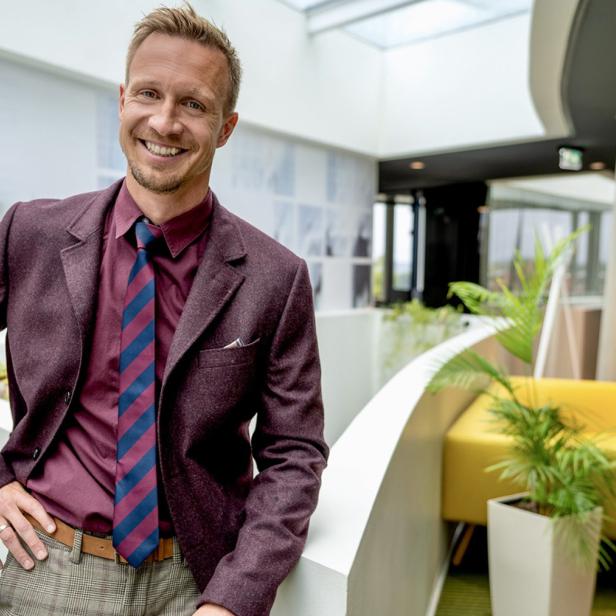 Mann im Anzug lächelt in modernem, hellen Büro mit Pflanzen und gelben Sitzmöbeln.