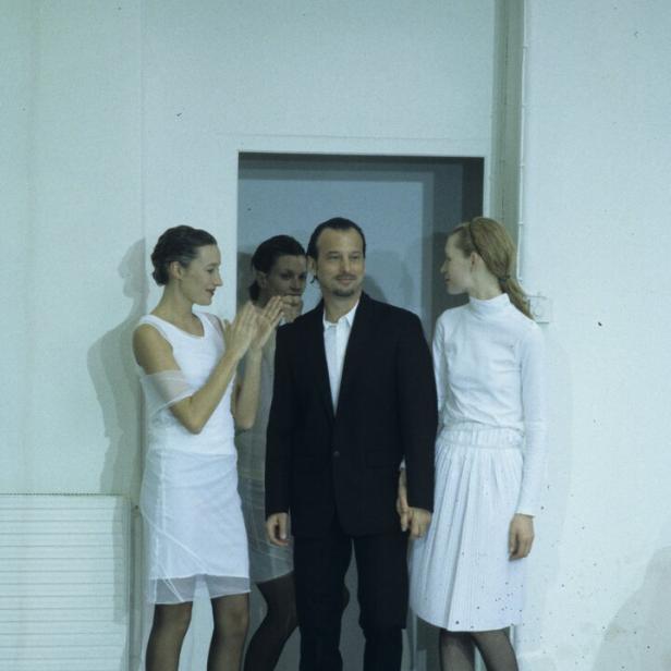 Mann im dunklen Anzug, umstellt von Frauen in weißen Kleidern