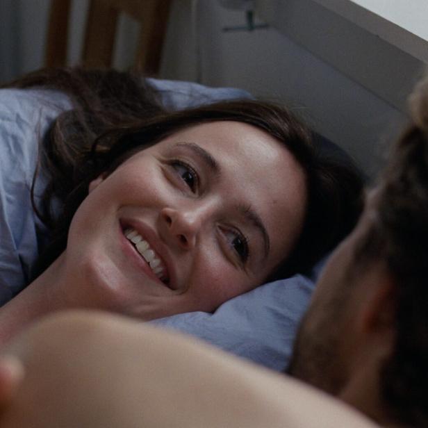 Lächelnde junge Frau liegt in einem Bett, blickt einen Mann an, den man nur von hinten sieht. 