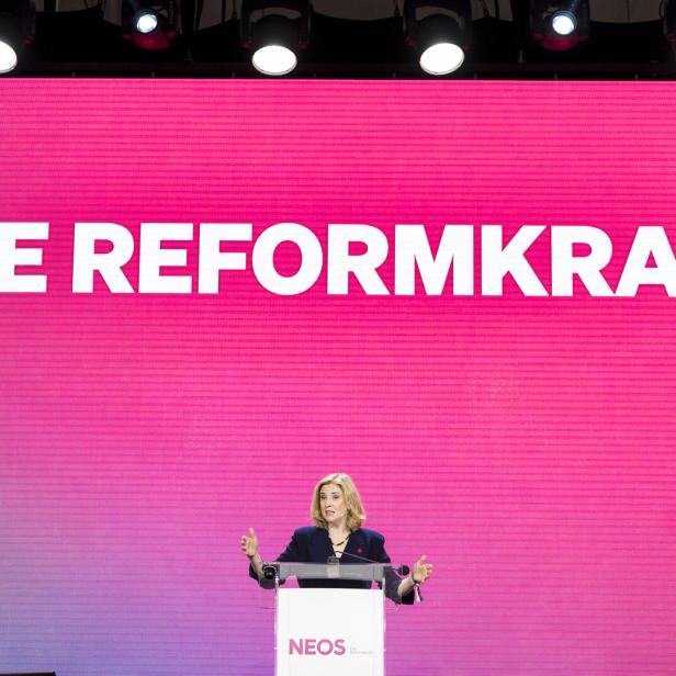Eine Frau, es ist Neos-Chefin Meinl-Reisinger, spricht am Rednerpult von NEOS vor einem pinkfarbenen Hintergrund mit der Aufschrift "DIE REFORMKRAFT".