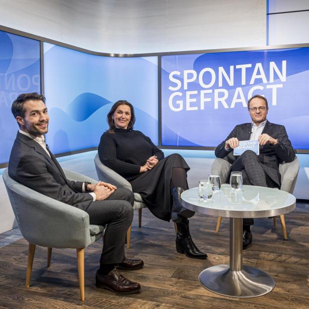 Drei Personen sitzen in Sesseln um einen runden Tisch mit Gläsern und Wasserkanne, im Hintergrund Bildschirm mit Schriftzug 'SPONTAN GEFRACT'.
