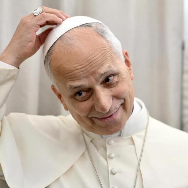 Der Papst in seinem weißen Ornat fasst sich an seine Mütze, während er den Kopf etwas schrägt hält.