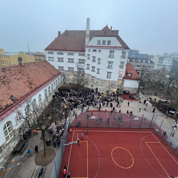 Viele Menschen versammeln sich auf einem Schulhof mit Sportplatz und historischen Gebäuden im Hintergrund.