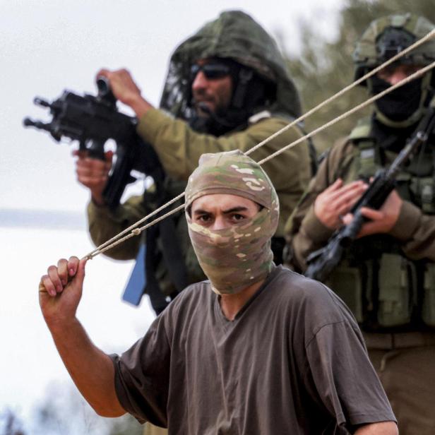 Ein israelischer Siedler steht vor zwei israelischen Soldaten, er lässt eine Steinschleuder über seinen Kopf kreisen.