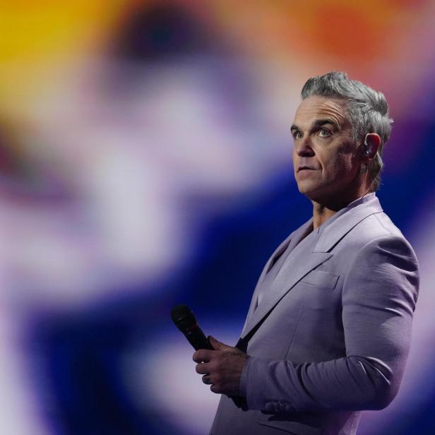 Robbie Williams im blasslila Anzug mit Mikro auf der Bühne