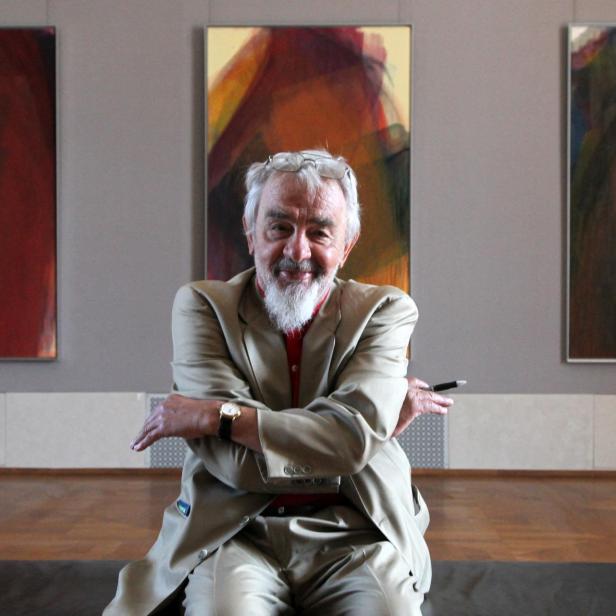 Der Künstler Arnulf Rainer, lachend, in hellem Anzug, hinter ihm drei seiner Übermalungs-Werke