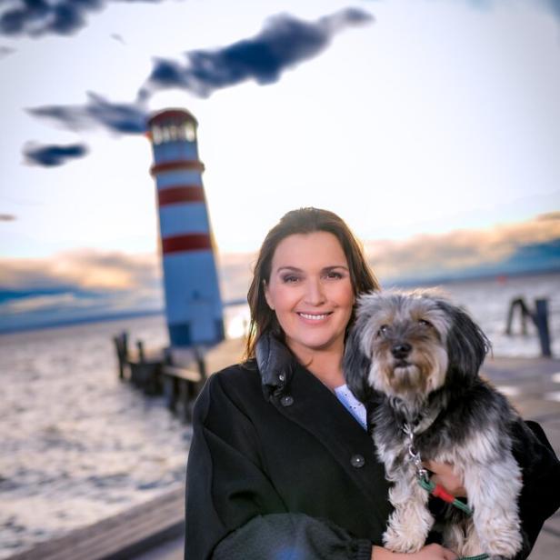 Barbara Karlich steht mit einem Hund im Arm vor einem Leuchtturm an einem See