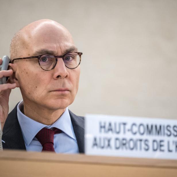 Volker Türk sitzt vor einem Schild ("haut-Commissaire aux droits de l'homme"), er hält sich ein Gerät ans Ohr und blickt in die Kamera.