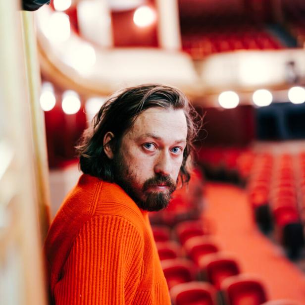 regisseur simon stone im parkett des burtheaters, mit rotem pullover und langen haaren