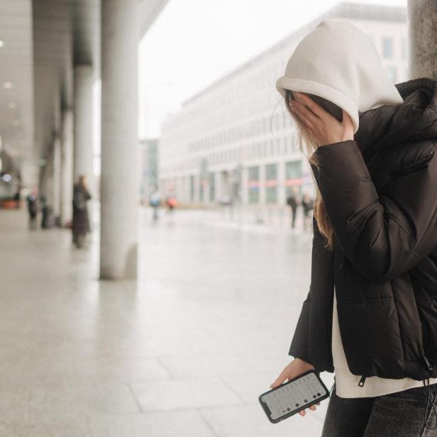 Ein Mädchen mit Kapuze über dem Kopf lehnt an einer Säule, das Smartphone und den Kopf hält.