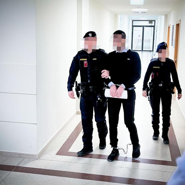 Ein Mann in Handschellen und Fußfesseln wird von zwei Polizisten durch einen Flur geführt.
