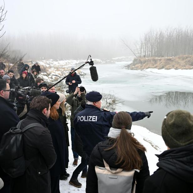 Eine Gruppe von Menschen steht am verschneiten Flussufer, ein Polizist zeigt auf das teilweise zugefrorene Wasser.