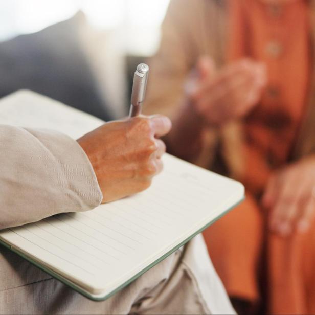 Eine weibliche Hand schreibt in ein leeres Notizbuch, gegenüber sitzt eine andere Frau, die erzählt. Eine Szene aus der Psychothterapie